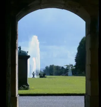The Emperor Fountain seen through a stone archway, with a tall water jet rising above a green lawn.