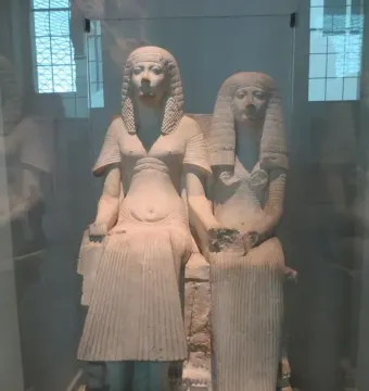 Horemheb and Amenia seated together on a shared throne, holding hands in a stone sculpture.
