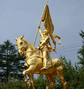 Golden equestrian statue of Joan of Arc in armor holding a flag-topped staff on a stone pedestal.