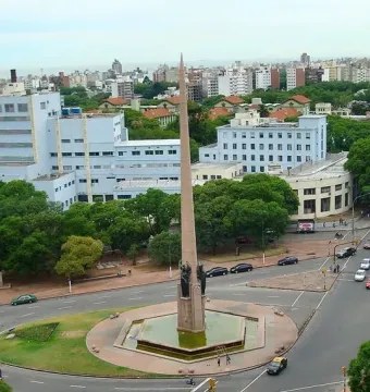 The Obelisk of Montevideo rises from a hexagonal water basin in the center of a busy traffic roundabout, with city buildings and trees around it.