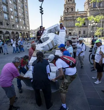 People gather in a sunlit public square around a wrapped structure being loaded onto a flatbed, with a cathedral tower in the background.