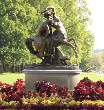 Bronze sculpture of Centauress and Faun on a stone pedestal in a park, with a manicured flower bed in front.