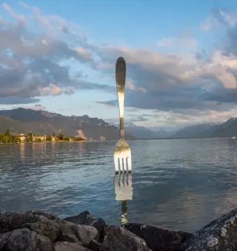 The Fork of Vevey sculpture stands in the lake with mountains and a cloudy sky in the background.