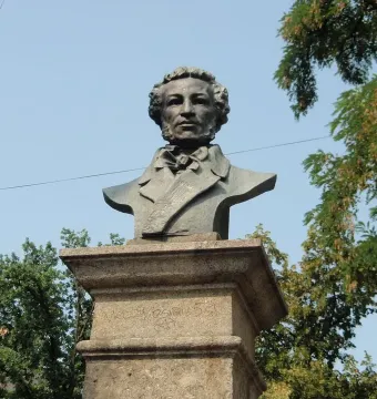 Dark bust of Pushkin on a stone pedestal outdoors, framed by trees against a blue sky.