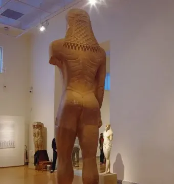 Rear view of the Sounion Kouros, a colossal ancient Greek stone statue of a nude male figure in a museum gallery.