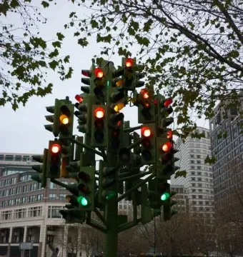 Traffic Light Tree sculpture made of green poles and many illuminated traffic lights in Canary Wharf, with buildings and vehicles behind it.