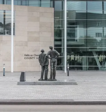 Two Working Men sculpture beside a modern glass-fronted building, with one figure standing taller and the other looking upward.