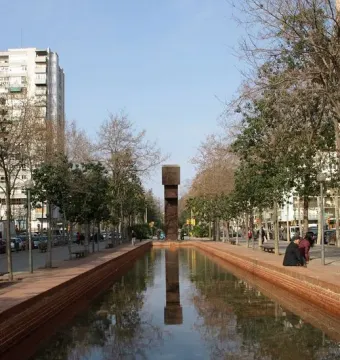 El Llarg Viatge at the end of a long reflecting pool, with trees, buildings, and people mirrored in the water.