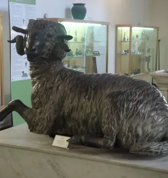 Bronze ram sculpture resting on a marble pedestal in a museum gallery.