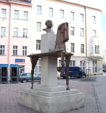 Bronze sculpture of Jaroslav Hašek with a fragmented horse on a multi-tiered stone pedestal in a paved square.