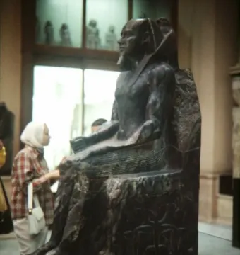 Khafre Enthroned seated dark pharaonic statue with a false beard in a museum, shown beside a person for scale.