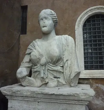 Madama Lucrezia, a weathered white marble statue of a woman in classical drapery on a stone pedestal beside a building wall and arched window.