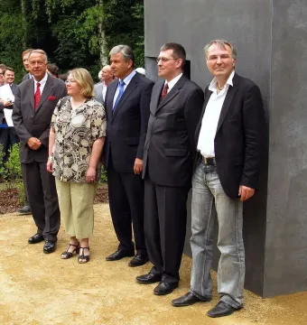 Five people stand beside the Memorial to Homosexuals Persecuted Under Nazism on a gravel path with trees and a crowd in the background.