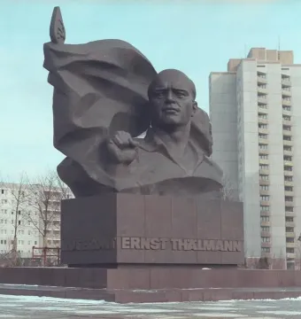 Ernst Thälmann Memorial bronze bust with a clenched fist and banner-like sculpture on a pedestal in a snowy urban setting.