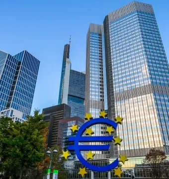 Blue Euro sculpture with yellow stars in front of modern glass skyscrapers.