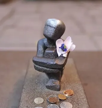 Järnpojke sculpture with a shiny head, a small flower in its chest, and coins scattered in front