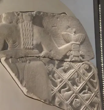 Fragmented relief from the Stele of the Vultures showing a large figure holding a staff with a bird emblem above smaller figures trapped in a net, with cuneiform text above.