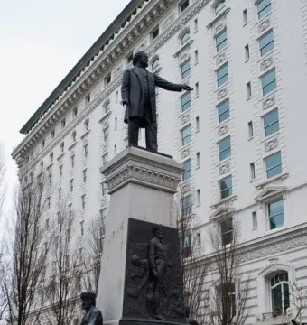 The Brigham Young Monument with a bronze figure atop a tall stone pedestal and relief sculptures at the base, with a white building and bare trees behind it.