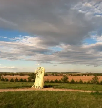 Carhenge sculpture with a pale vintage car standing vertically in a grassy field under a cloudy sky.
