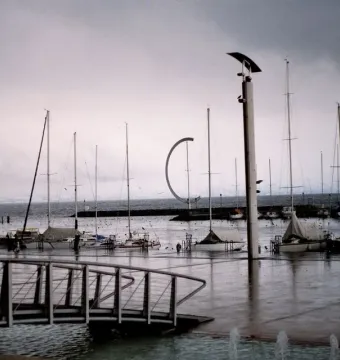 Éole, a dark C-shaped metal sculpture, stands on a breakwater near sailboats in the marina.