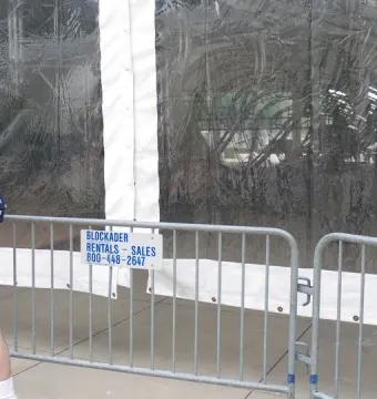 A person in a patriotic shirt and top hat stands behind a metal barricade looking toward the Burnham Pavilions inside a clear tent.