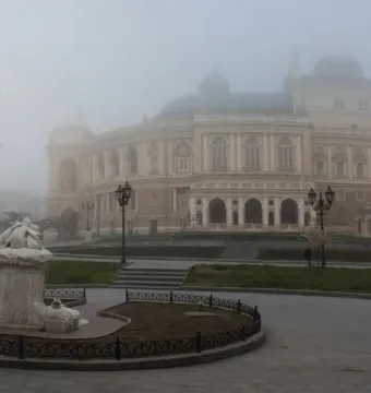 A light-colored stone Children and Frog monument stands in a fenced oval plot on a foggy square, with an ornate building in the background.
