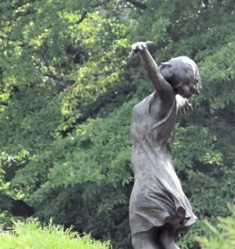 The Dancing Girl sculpture in dark metal, seen from a low angle among blurred green foliage.