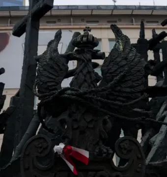 Dark metal memorial sculpture with a crowned eagle emblem, a large cross, and a ribbon tied at the base.