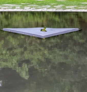 A dark triangular slab of the Memorial to the Murdered Sinti and Roma of Europe floats on water with a small cluster of flowers at its center.