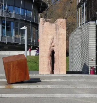Pi-Chacán stone sculpture with a deep vertical crevice, standing beside a rusted rectangular block on a paved area.