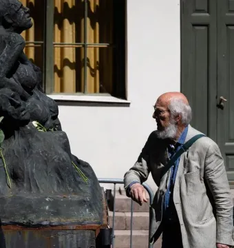 Pillar of Shame sculpture with white roses at its base, flanked by two men observing it