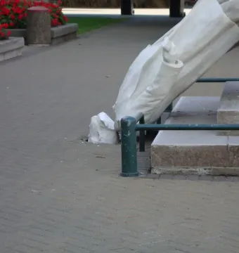 Rolleston Statue toppled over on a paved walkway beside a stone base, with red flowers and a green railing in the background.