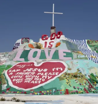 Salvation Mountain covered in colorful religious murals, with a white cross on top and large painted words including LOVE and GOD IS.