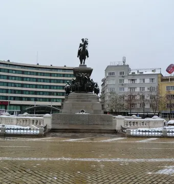The Tsar Liberator Monument stands on a stone pedestal in a snowy Sofia square, with buildings and a curved modern structure behind it.