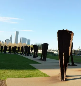 Agora sculpture with dozens of rusted, headless human-like figures spread across a grassy area and walkway in front of the Chicago skyline.