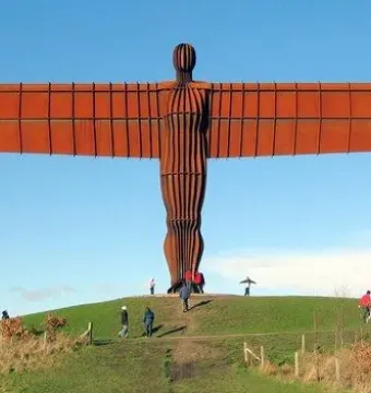 The Angel of the North sculpture stands on a green hill with people walking near its base under a clear blue sky.