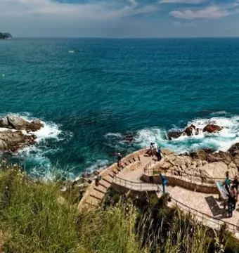 Dona Marinera bronze statue on a stone lookout above the sea and rocky coastline, with people walking nearby.