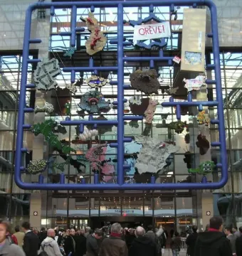 Entropa installation with a large blue grid frame and colorful attached shapes in a modern interior, with people gathered below.