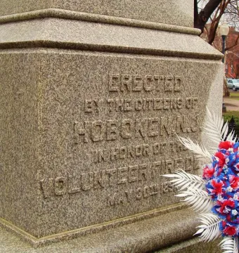 Firemen\'s Monument stone pedestal inscription honoring the volunteer fire department, with a red, white, and blue floral wreath beside it.