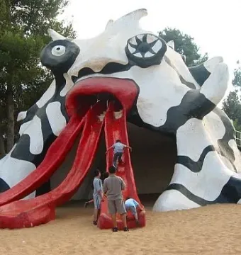The Golem monster sculpture with three red slide-like tongues, with children climbing on it in a sandy park area.
