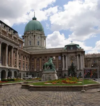 King Matthias Fountain sculpture group in a cobbled courtyard beside flowerbeds and a grand building with a green dome.
