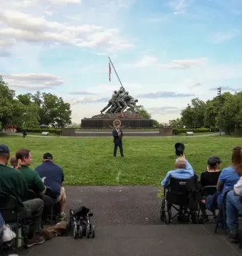 The Marine Corps War Memorial stands in a grassy park as a crowd sits facing a brass performer in the foreground.