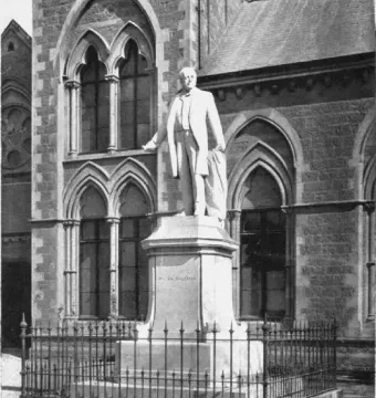 The Rolleston statue stands on a tall hexagonal pedestal behind a dark iron fence, with a stone building in the background.