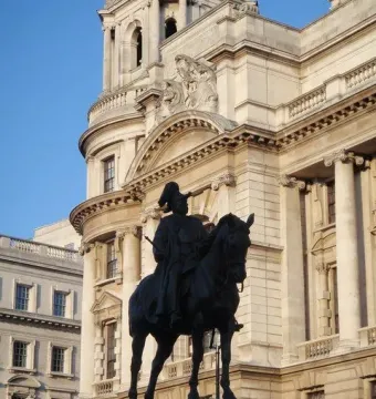 The Duke of Cambridge equestrian statue silhouetted against a grand stone building and blue sky.