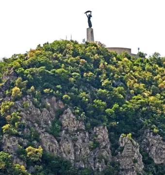 The Liberty Statue stands on a tall pedestal atop a wooded hill against a bright sky.