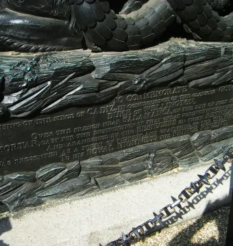 Close-up of the Cádiz Memorial plaque set into a sculpted base with textured relief and a dark metal post beside it.