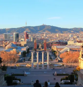 The Four Columns standing in a plaza with a circular fountain in front and a city and mountains in the distance.
