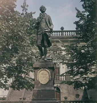 Goethe Monument on a tall granite pedestal with a golden plaque, surrounded by trees and a grand building in the background.