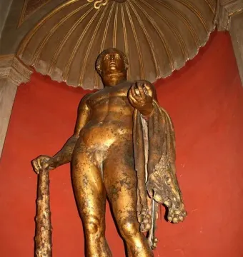 Hercules statue holding a club, with a lion skin and apples, set against a red wall beneath a gilded alcove.