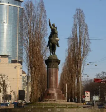 The Monument to Mykola Shchors, a bronze equestrian statue on a tall stone pedestal along a city street with trees and buildings nearby.
